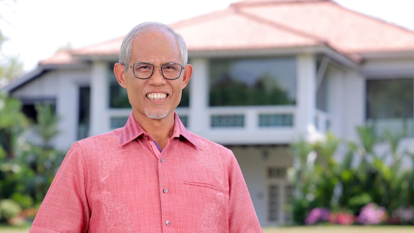 Man in pink shirt; white building with terracotta roof behind.